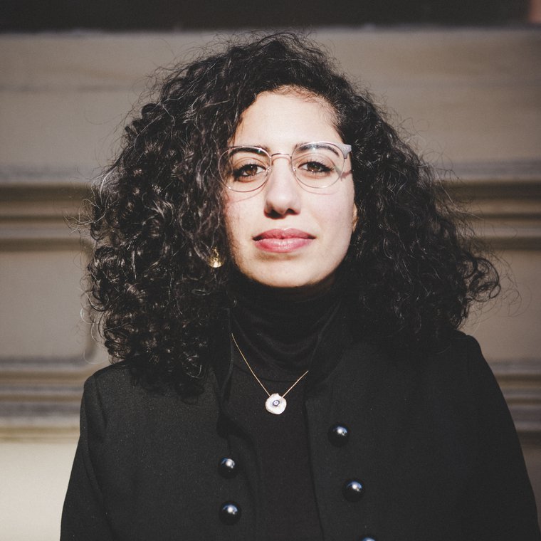 Image of Middle Eastern woman sitting in middle of the frame wearing black, with curly hair down to her shoulders