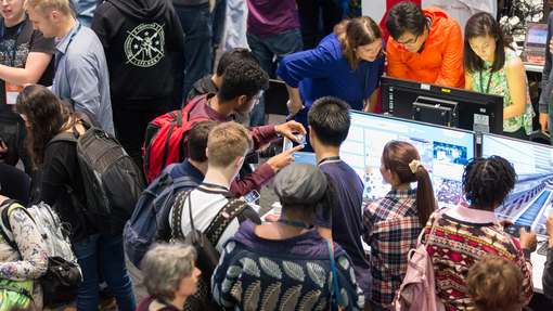 Crowd of people gather around a table display with screens at a busy indoor exhibition. Some interact with the display while others observe.