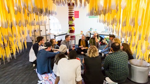 People sit in a circle on the floor under hanging yellow strips of fabric in a brightly lit room with soft seating and posters on the wall.