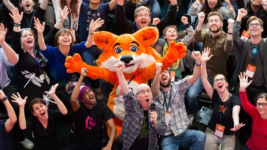 Group of people cheer with raised arms around a person in a large orange fox mascot costume at a crowded indoor event.