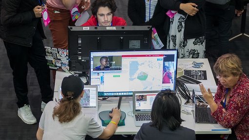 A group of people gathers around a desk with multiple monitors and laptops displaying maps and video calls.