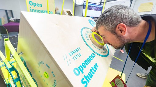 A person leans forward to look through a circular viewer labeled "Openness Shutter" on a wooden interactive exhibit.