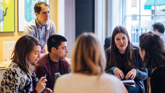 Seven people gather around a table indoors. One person stands, while the others sit with notebooks and papers on the table.