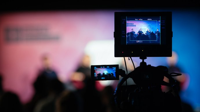 A camera on a tripod records a panel discussion, with the stage visible on the camera screens and blurred audience in the foreground.