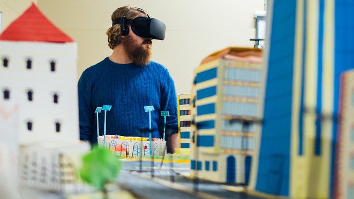 Person wearing a virtual reality headset stands behind a colorful scale model of a city with paper buildings, streets, and trees.