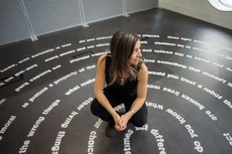 An artist, sitting with one of their art pieces, of various moral questions in the shape of a labyrinth on the floor.