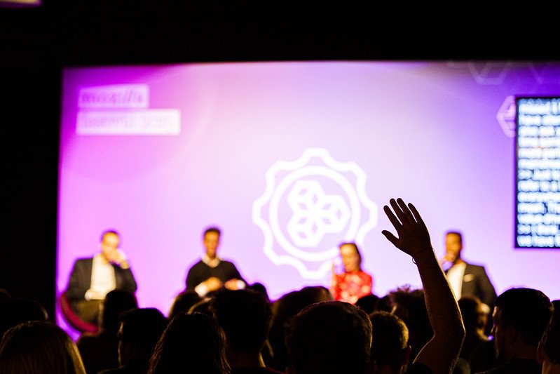 A group of 4 panelists sitting on a stage with a purple background. In the fore ground, you can see the backs of people's heads with one hand raised.