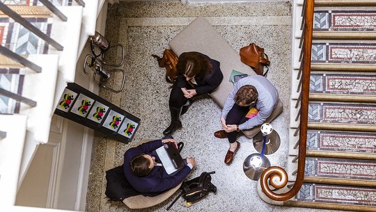 Overhead view of three people sitting in a stairwell lounge area, talking. One holds a laptop, and bags are on the floor and bench nearby.