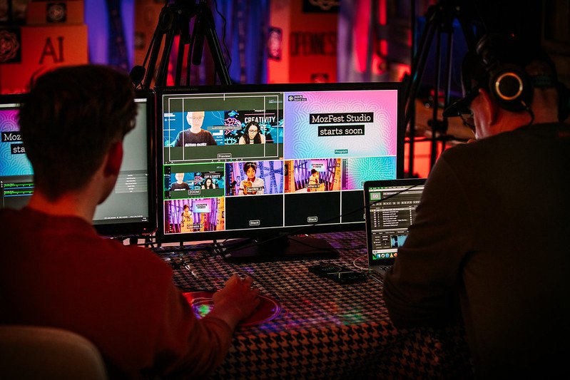 A behind the scenes photograph showing two people working with broadcast equipment, with a monitor in the middle that shows different cameras and graphics, including a presenter at a lectern, a video conference with two people side by side and a graphic that reads MozFest Studio starts soon.