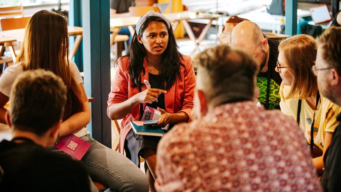 A group of people sit in a circle indoors, with one person in a red jacket speaking while others listen attentively.