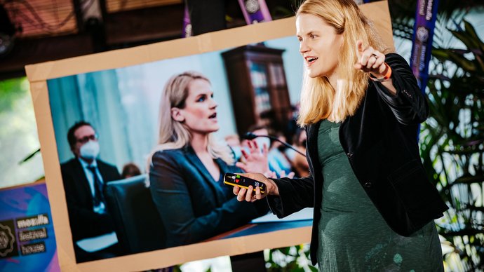 Frances Haugen stands and gestures while speaking beside a large screen displaying an image of her speaking at a committee..