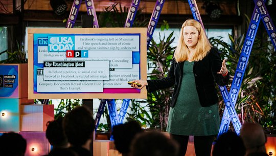 Frances Haugen s’exprime sur la scène du Mozilla Festival, faisant des gestes tout en présentant une diapositive avec les titres des journaux sur l’impact des réseaux sociaux.