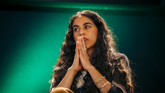 Mona Chalabi sits with her hands pressed together against a green-lit background.