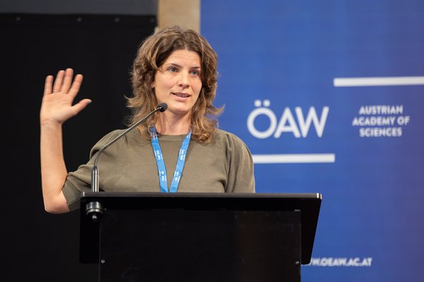 Woman with blue lanyard speaks in front of a microphone at the Austrian Academy of Sciences