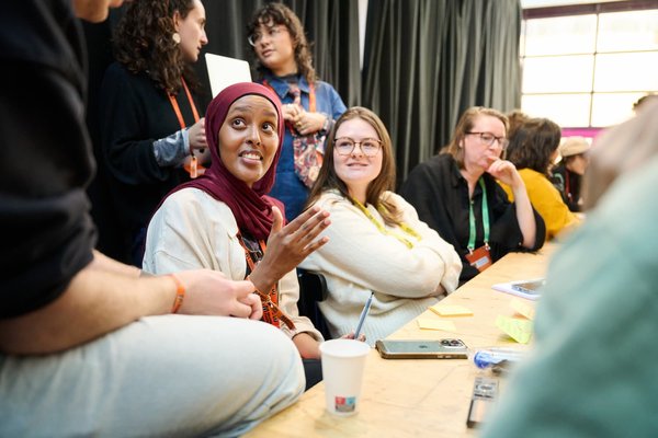 A person in a maroon hijab gestures while speaking at a conference table; others around the table watch, one smiling with arms crossed, lanyards and sticky notes nearby.