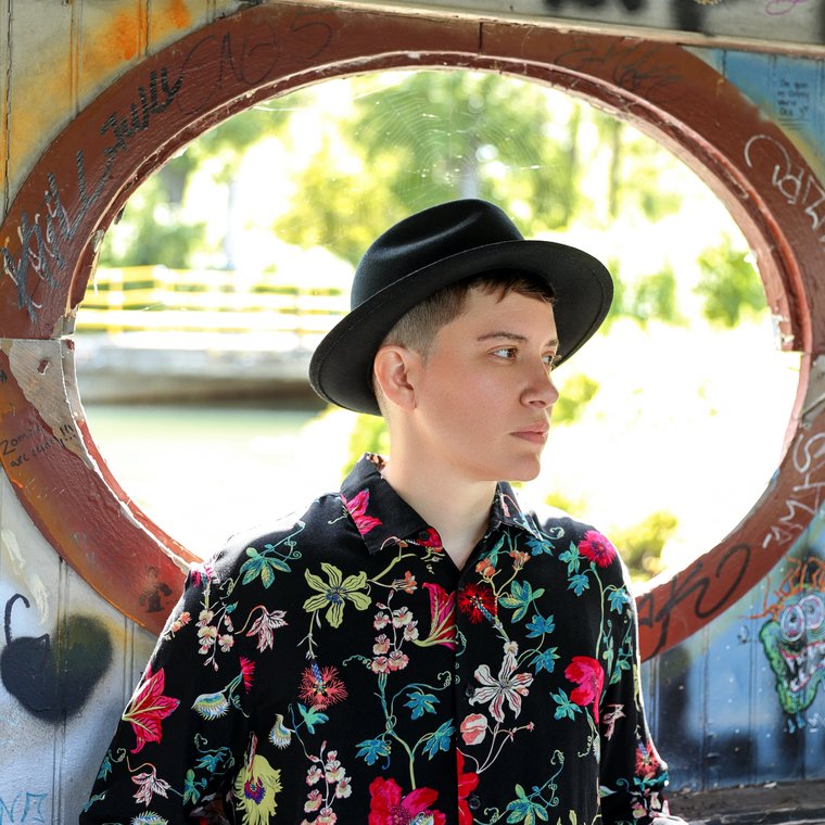 A person looking to the right, wearing a black brimmed hat and colorful shirt. An oval window is in the background.