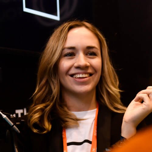 A person with medium length brown hair wearing a black and white shirt with orange accents smiling and looking off into the distance. One hand is slightly raised.