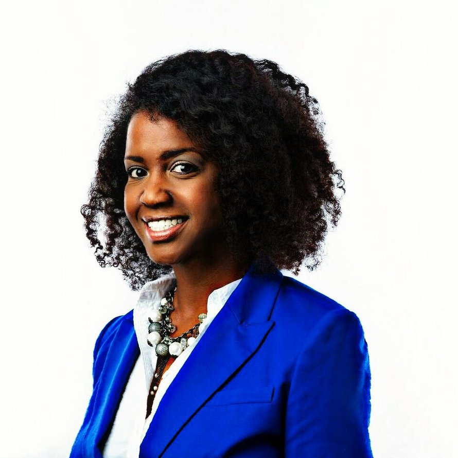 A person with shoulder length dark curly hair wearing a white buttoned shirt and blue blazer smiling at the camera