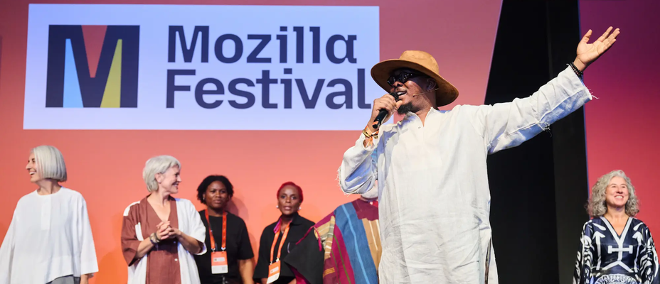 Malik Afegbua speaks into a microphone with one arm raised on stage. People in varied outfits stand behind him.