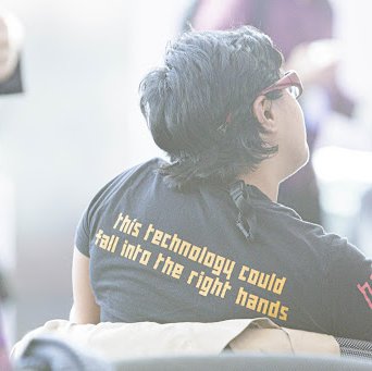 una persona con anteojos de espaldas a la cámara, su camiseta azul tiene una escritura amarilla en la parte posterior que dice "esta tecnología podría caer en las manos adecuadas"