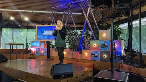 A wooden stage with large windows in the background. A person is standing, holding a microphone, and speaking into it.