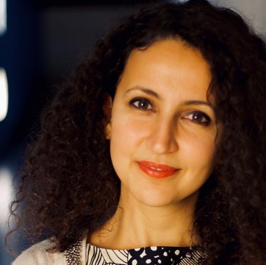 A person with medium length dark curly hair leaning against a wall and looking at the camera