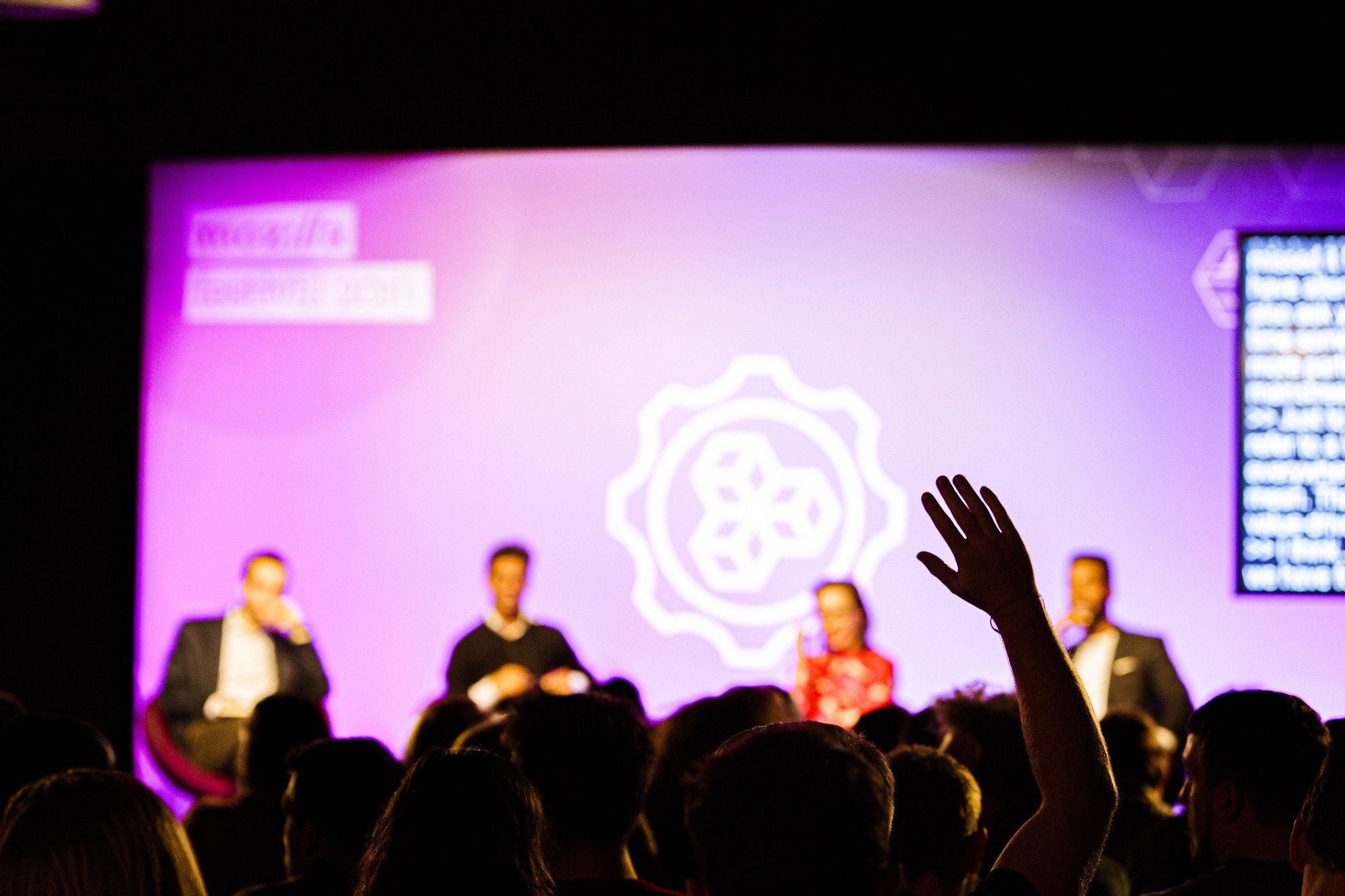 A panel of four speakers on stage in the background, with a person raising their hand in the foreground.