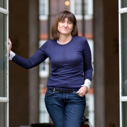 A women looking at the camera with one hand in her pocket and another leaning against a wall, wearing Blue long sleeve shirt and blue jeans