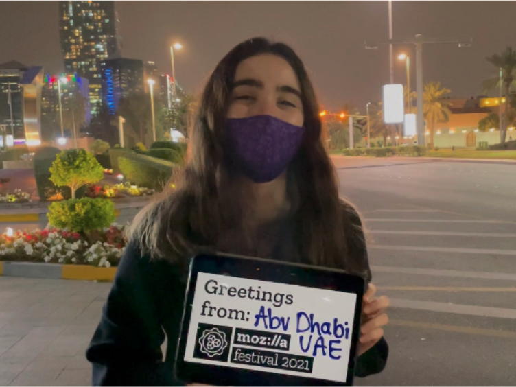 Person with long, brown hair and wearing a purple face mask looking at the camera and holding a white sign that says "Greetings from Abu Dhabi, UAE, Mozilla Festival 2022