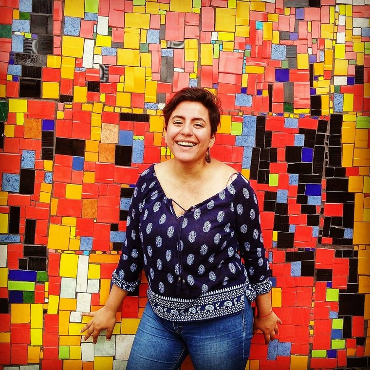 Person standing in front of colorful background. Person has short dark hair, is smiling at the camera, and is wearing a patterned shirt and blue jeans.