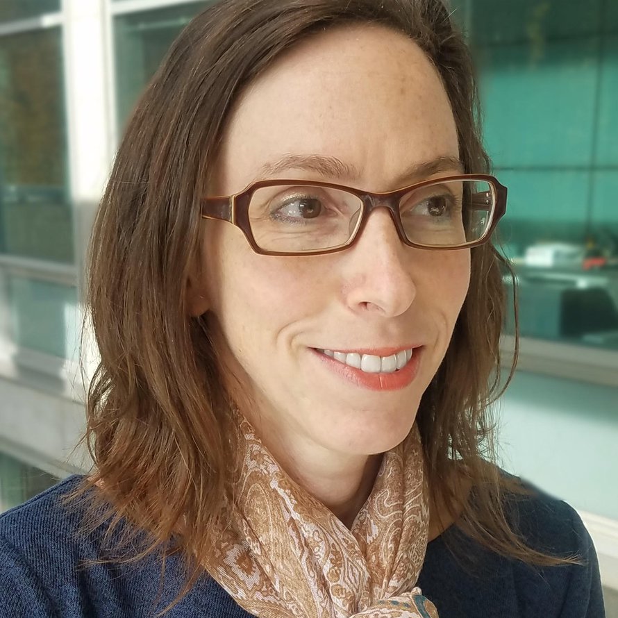 A person with medium length brown hair wearing dark rimmed glasses, a blue shirt, and light colored scarf smiling into the distance