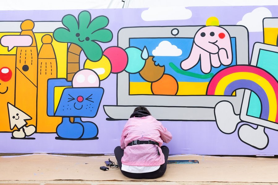 An artist wearing a pink shirt sits in on the ground in front a painted mural they are working on