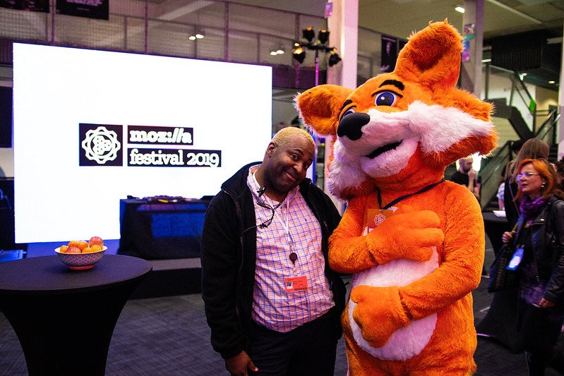 MozFest Volunteer in black sweater and plaid shirt standing next to person in foxy costume, smiling at the camera