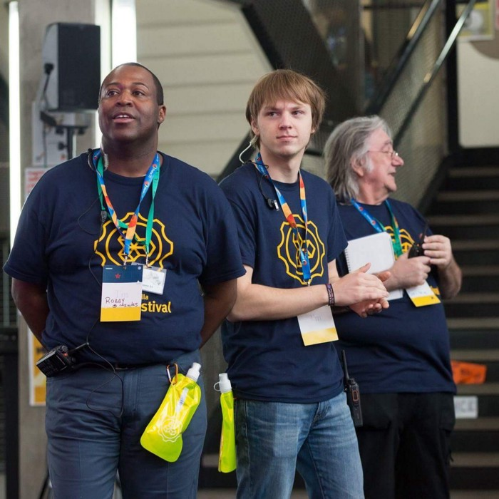 Three volunteers at MozFest wearing blue festival shirts and name tag lanyards.