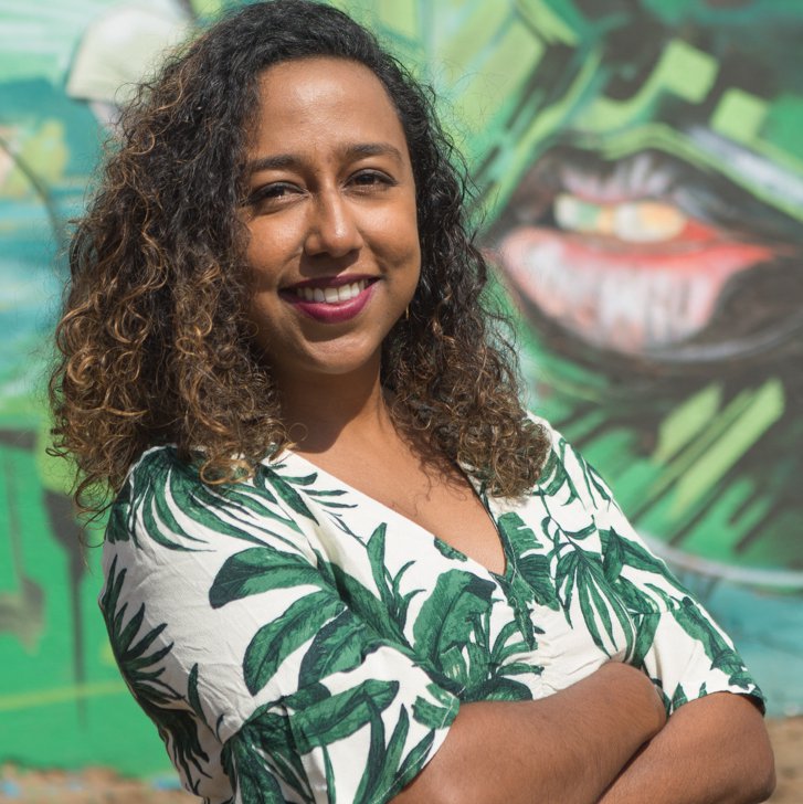 A person with arms folded, brown curly hair, a tropical shirt smiling at the camera.Posed against a tropic background