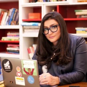 Person with long, dark hair and dark rimmed glasses leaning against a desk with a laptop open, looking at the camera.