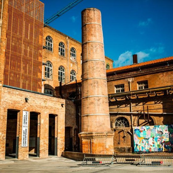 Fabra i Coats: brick industrial buildings with a tall chimney and graffiti, under a blue sky.