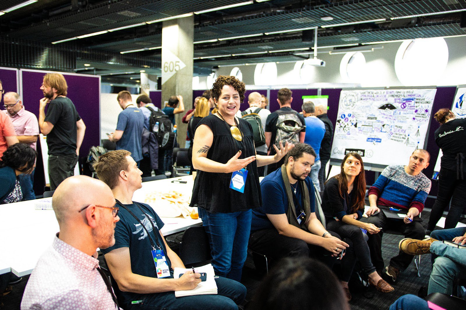 Person standing up and speaking amidst a room full of people sitting in chairs. Background has posters on temporary walls and people chatting with one another.