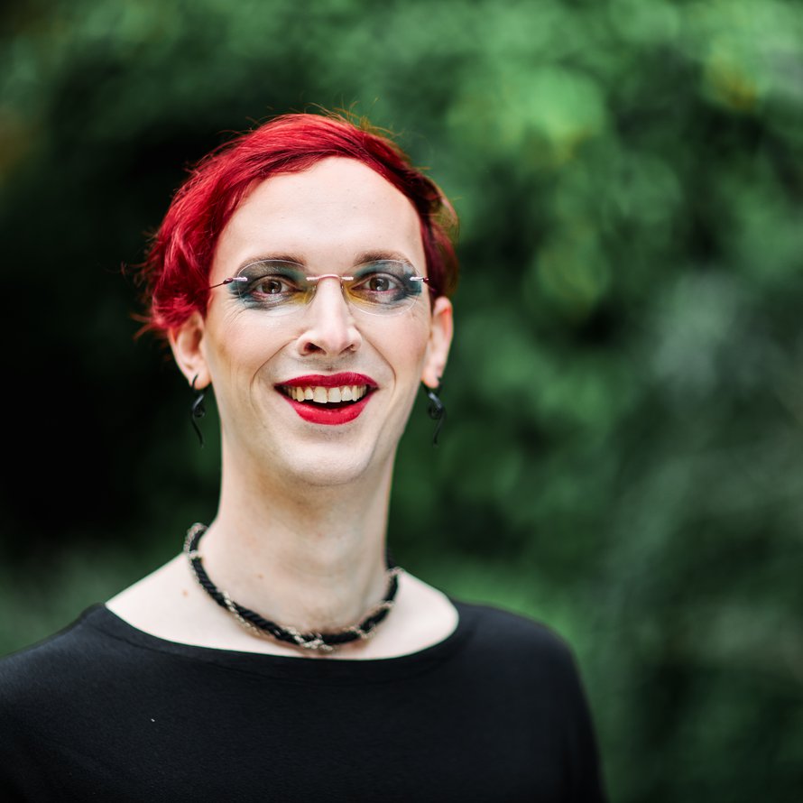 A person with red, short hair wearing a black shirt and necklace smiling at the camera