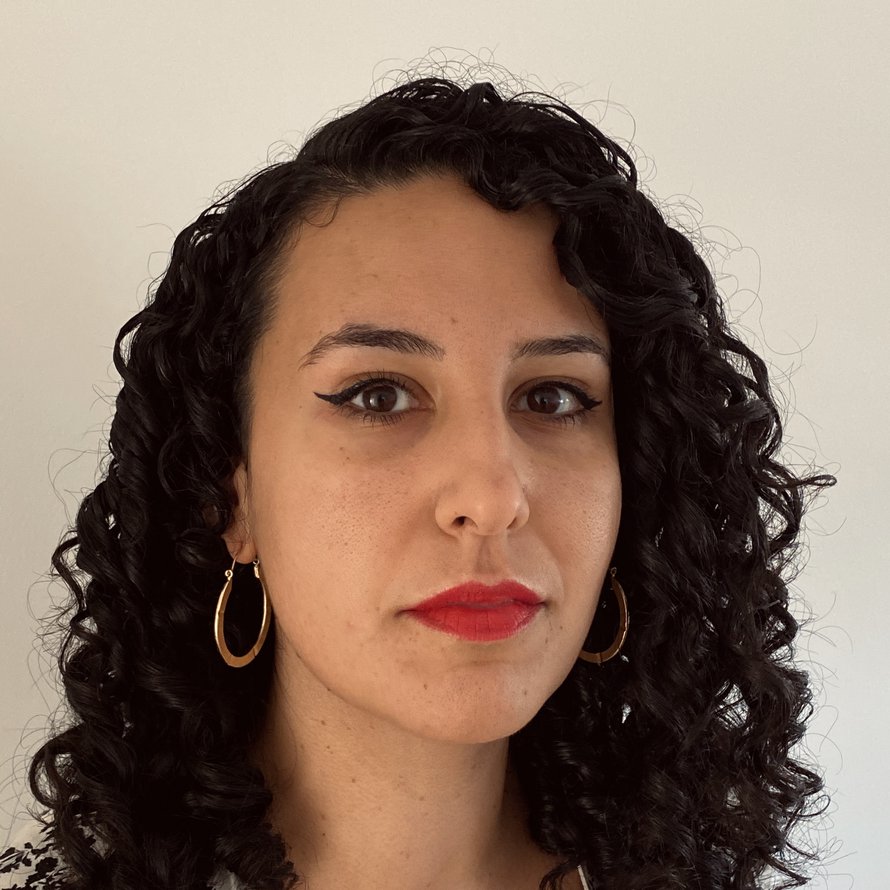 A person with dark curly hair, wearing hoop earrings and a collared shirt looking at the camera