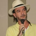 A person wearing a white rimmed hat with a black ribbon, holding a microphone and wearing a yellow polo shirt.