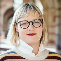 A person with medium length blond hair and bangs, wearing dark rimmed glasses and a striped sweater, smiling at the camera