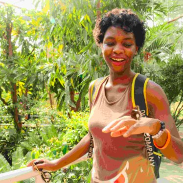 Person with dark, short, curly hair standing with a natural setting in the background, holding their hand up and a butterfly sitting on it.
