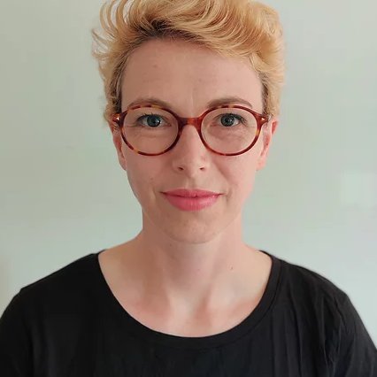 A person with short blond hair wearing red rimmed glassed and a black shirt looking at the camera