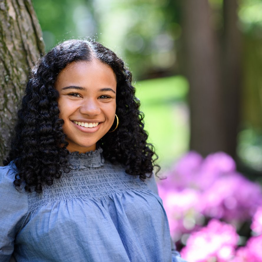 Person with long, dark, curly hair leaning against a tree and smiling at the camera