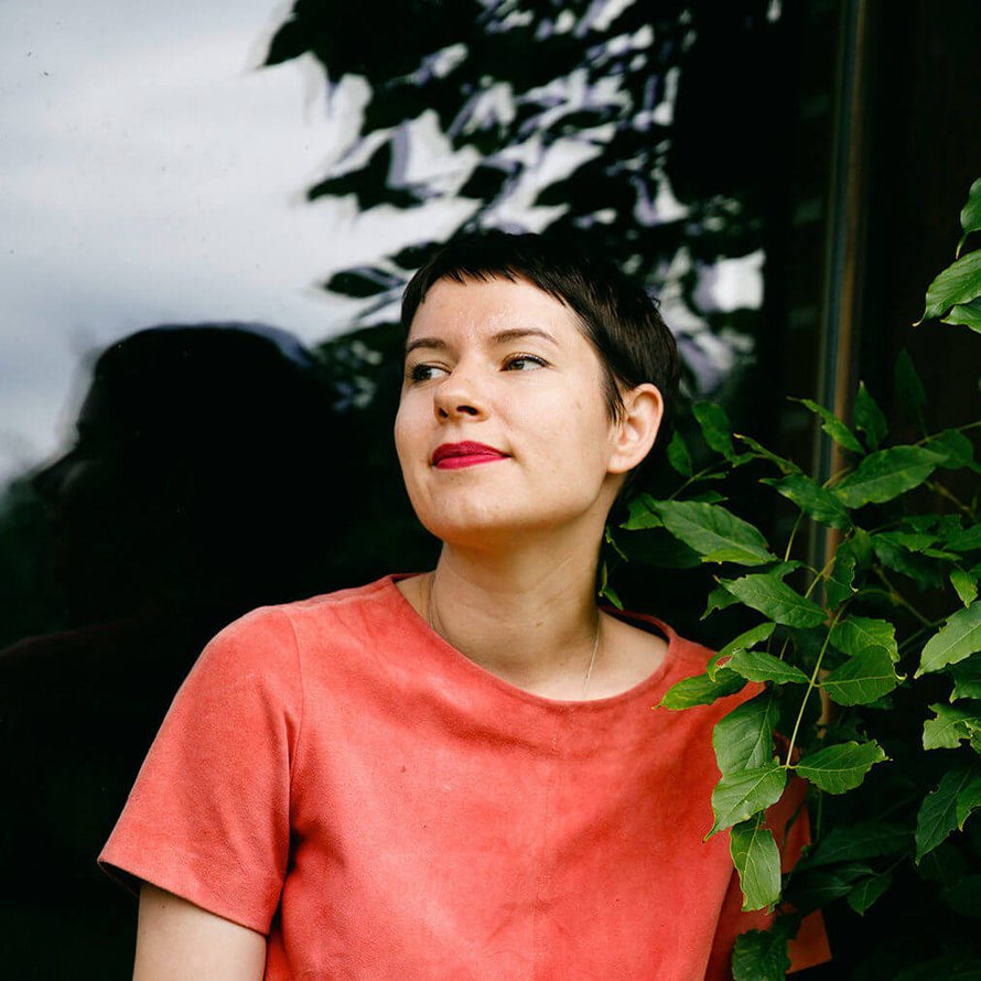 A person with short dark hair wearing a pink short sleeved shirt standing next to a plant and looking off into the distance