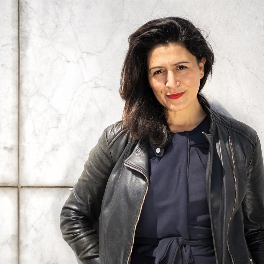 A person with long dark hair wearing a dark shirt and black jacket leaning against a white wall