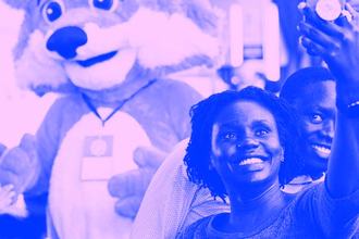 Three women stand in front of the Firefox mascot, Foxy, and take a selfie with him while other MozFest attendees cross in the background.