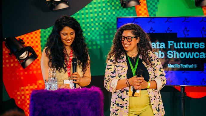 Two people stand on stage with microphones, speaking in front of a colorful backdrop and a screen displaying "Data Futures Lab Showcase."