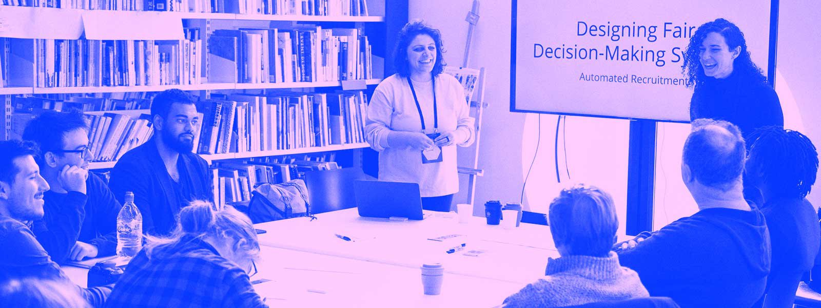 Two facilitators stand at the head of a conference table where several people sit in a library setting and a large monitor that says "Designing Fair Decision-Making Systems."